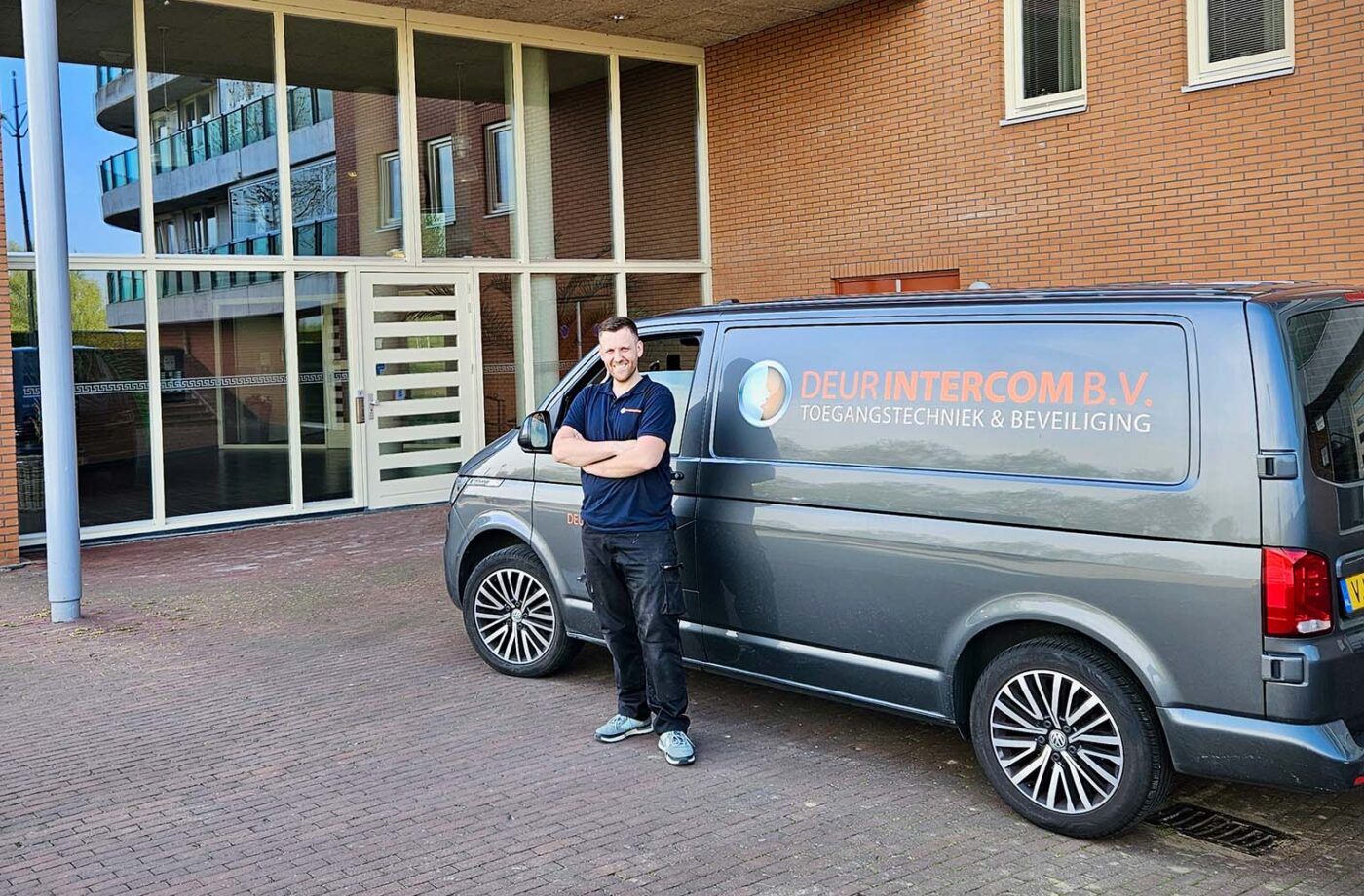 Monteur van Deurintercom B.V. staat met gekruiste armen naast een bedrijfsbus met logo “Deurintercom B.V. – Toegangstechniek & Beveiliging”, geparkeerd voor de entree van een modern appartementencomplex.