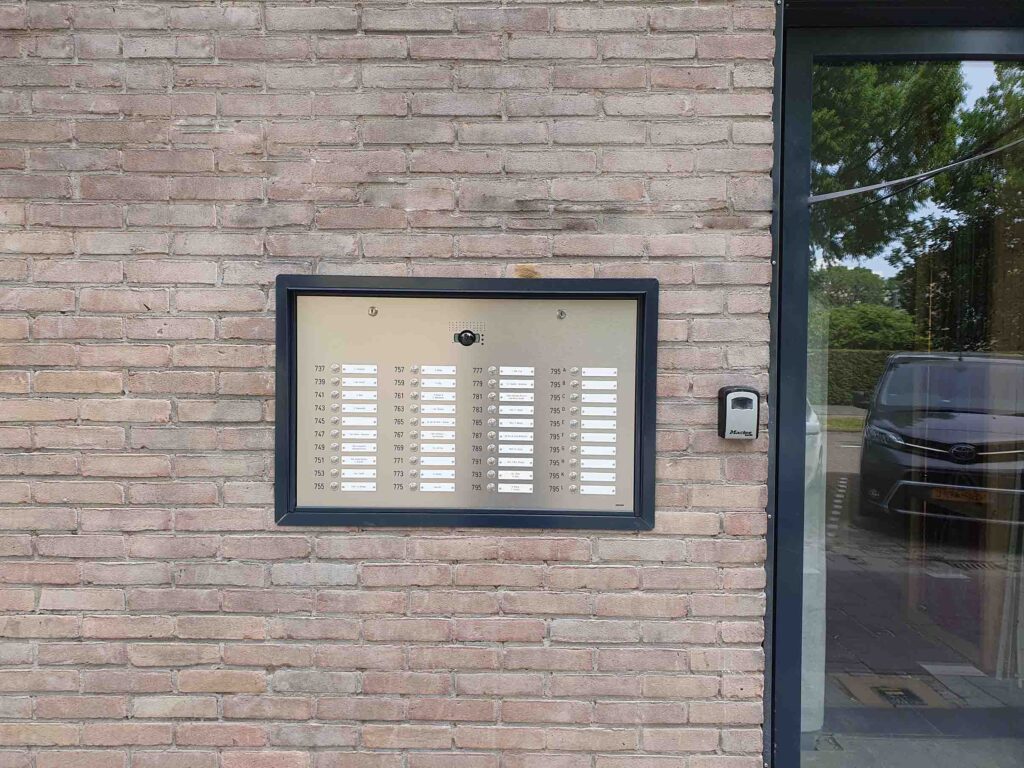 Nieuw RVS belpaneel met geïntegreerde camera en meerdere naamplaatjes, gemonteerd in de gevel naast de entree van een appartementencomplex.